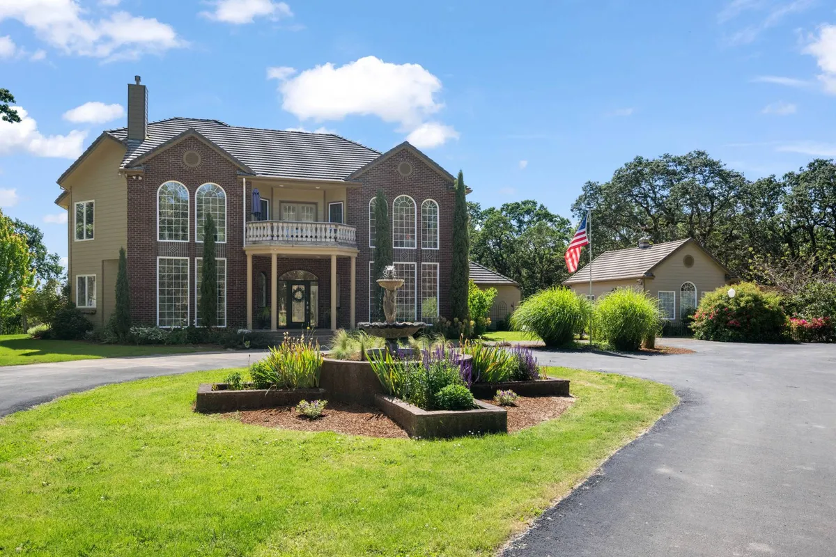 A grand exterior shot showing the estate’s main entrance, a central fountain, beautifully landscaped gardens, and a welcoming front porch area. The setting is serene and inviting, suggesting memorable arrivals and outdoor gatherings.