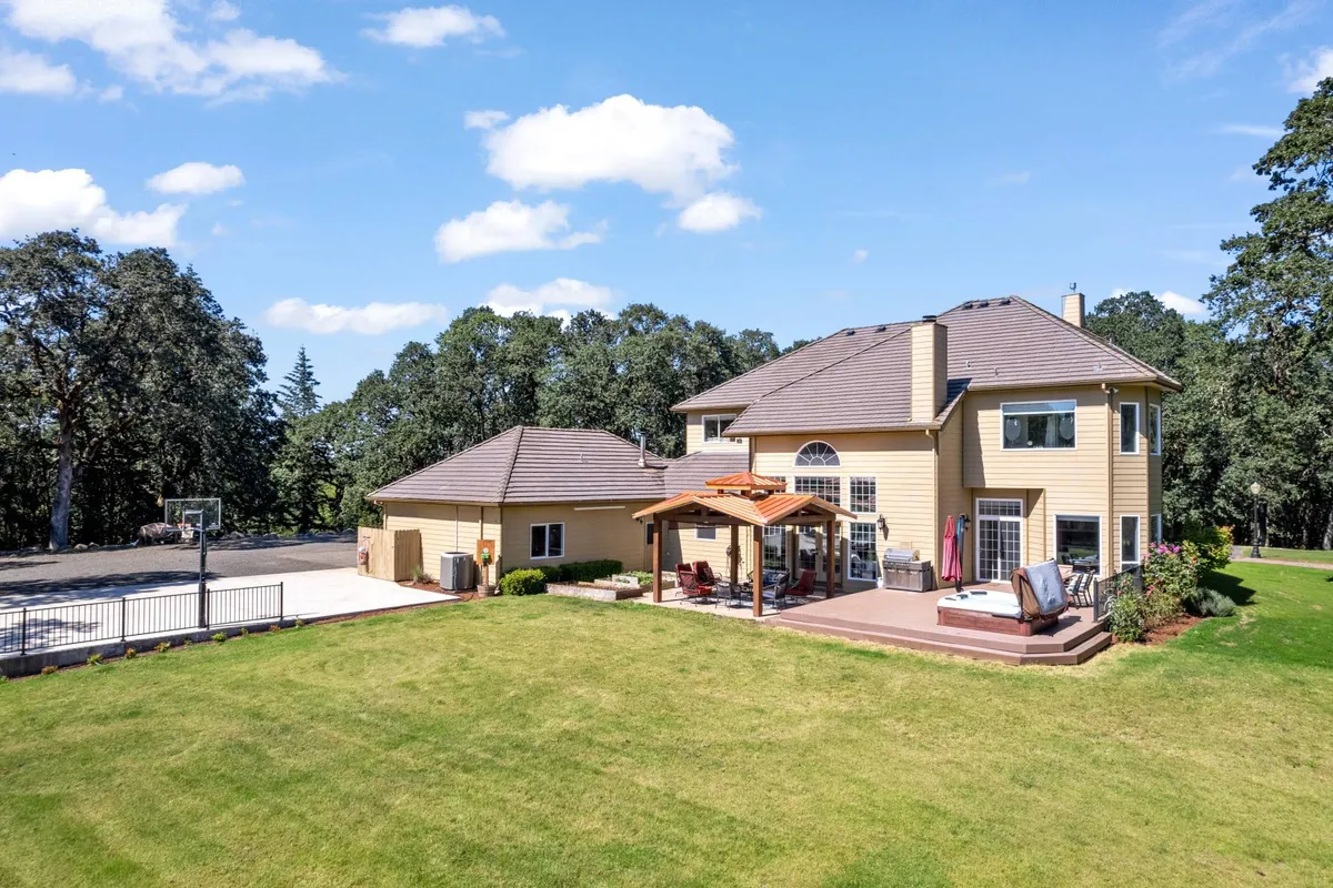 This exterior image showcases the main house with its welcoming deck area, adjacent basketball court, and the lush oak forest backdrop. The spacious setting is ideal for outdoor family gatherings, activities, and recreation, highlighting the estate's generous grounds and amenities.