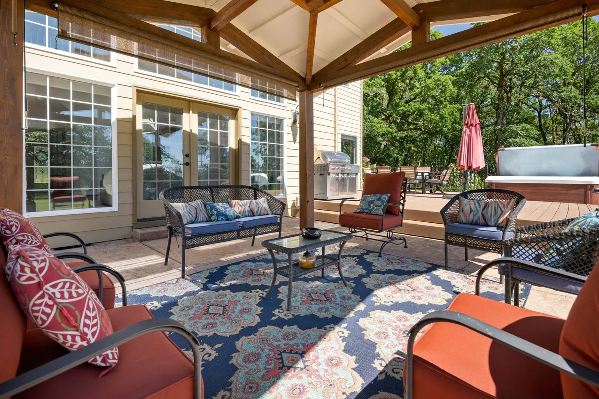 An inviting outdoor lounge area featuring a comfortable seating arrangement with colorful cushions and a decorative rug, set beside a hot tub and an outdoor grill. The area is designed for relaxation and socializing, with the fire pit and lush greenery in the background hinting at the estate's expansive natural setting.