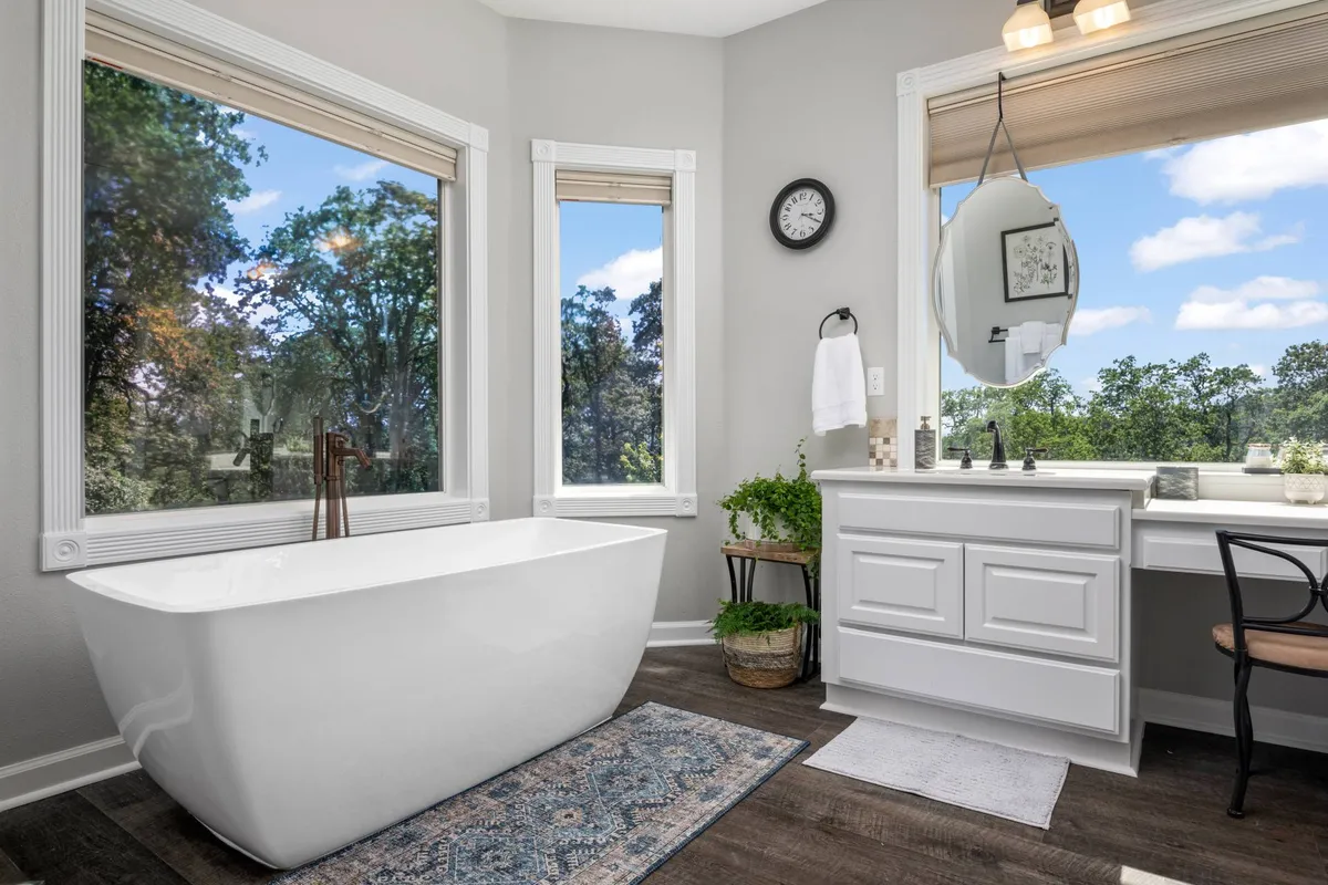 A spa-inspired bathroom with a freestanding bathtub, large windows looking onto greenery, a vanity area, and decorative plants. The scene feels bright, refreshing, and luxurious—perfect for relaxation and self-care.