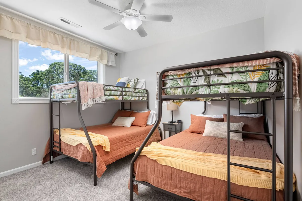 This image features a cheerful bunk room with bunk beds dressed in colorful bedding and decorative pillows. Large windows fill the space with natural light, and a nightstand with a lamp adds functional coziness. The space feels playful yet restful, ideal for children or group stays.