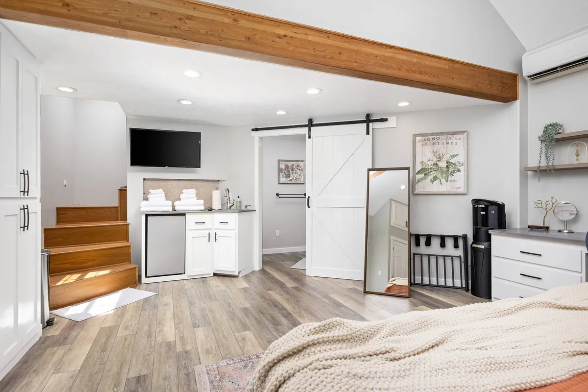 A calm, inviting guest suite featuring a comfortable bed with a knitted throw, a sliding barn door, a kitchenette area, mirror, and a staircase leading to a loft above. The space feels self-contained and perfect for a private guest experience.