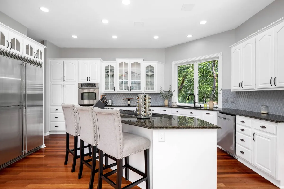 A modern and welcoming kitchen with an island bar and stools, stainless steel appliances, granite countertops, and large windows that overlook surrounding greenery. The open layout encourages group interaction, cooking, and casual dining.