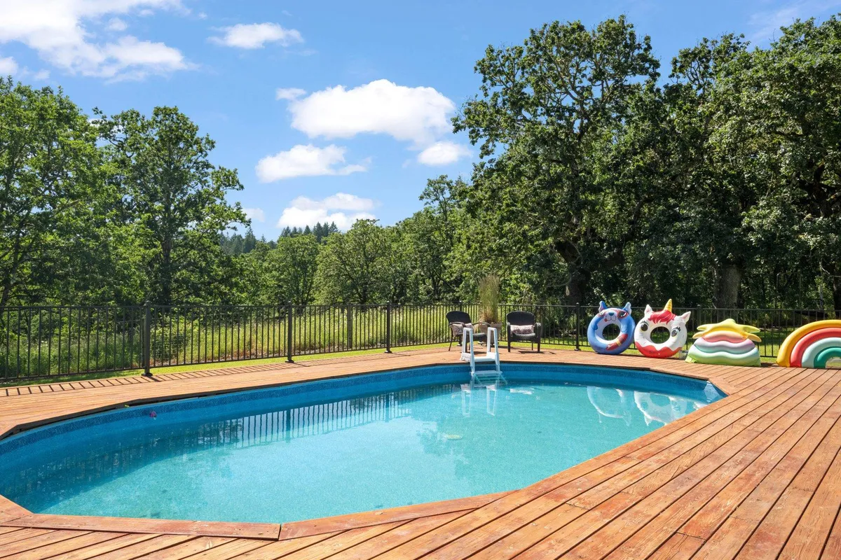 The image features a vibrant pool area with a wooden deck, colorful floats, and lush greenery beyond. The inviting pool is ideal for summer relaxation, group gatherings, and playful family moments under the sun.