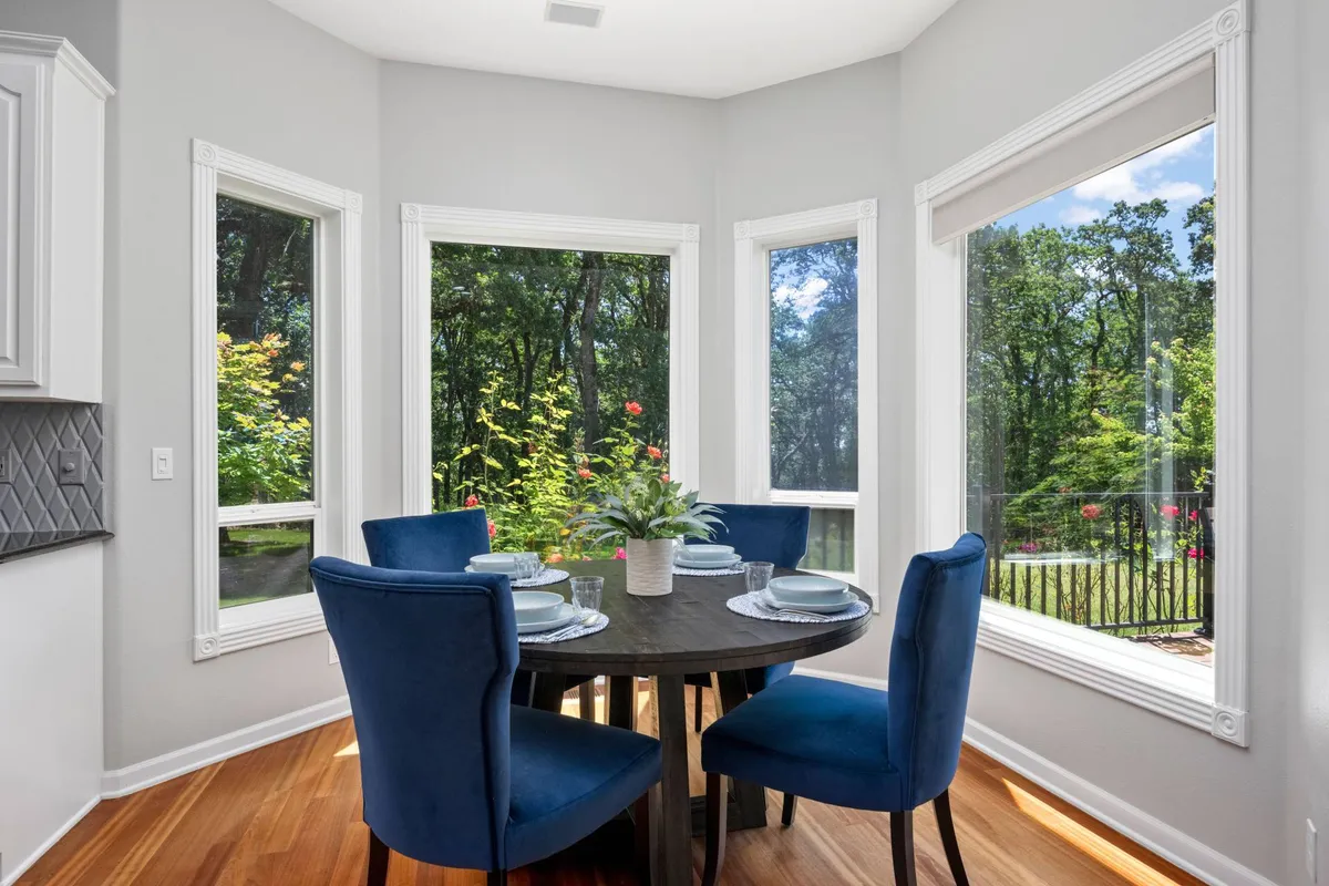 A sunlit dining area with large windows that frame scenic greenery, blue upholstered chairs around a round table, and a floral centerpiece. The bright, airy atmosphere highlights both comfort and the proximity to the outdoor landscape.