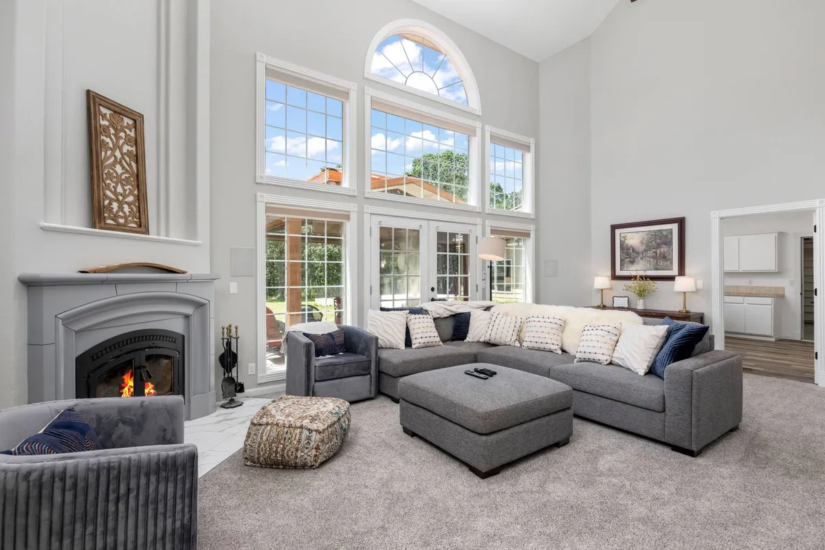 This image showcases a spacious living room featuring a large sectional sofa, a prominent fireplace, and expansive windows that fill the room with natural light and provide views of the surrounding Savannah oaks. Decorative wall art and plush seating create a cozy and inviting atmosphere, perfect for family gatherings, movie nights, and entertaining guests.