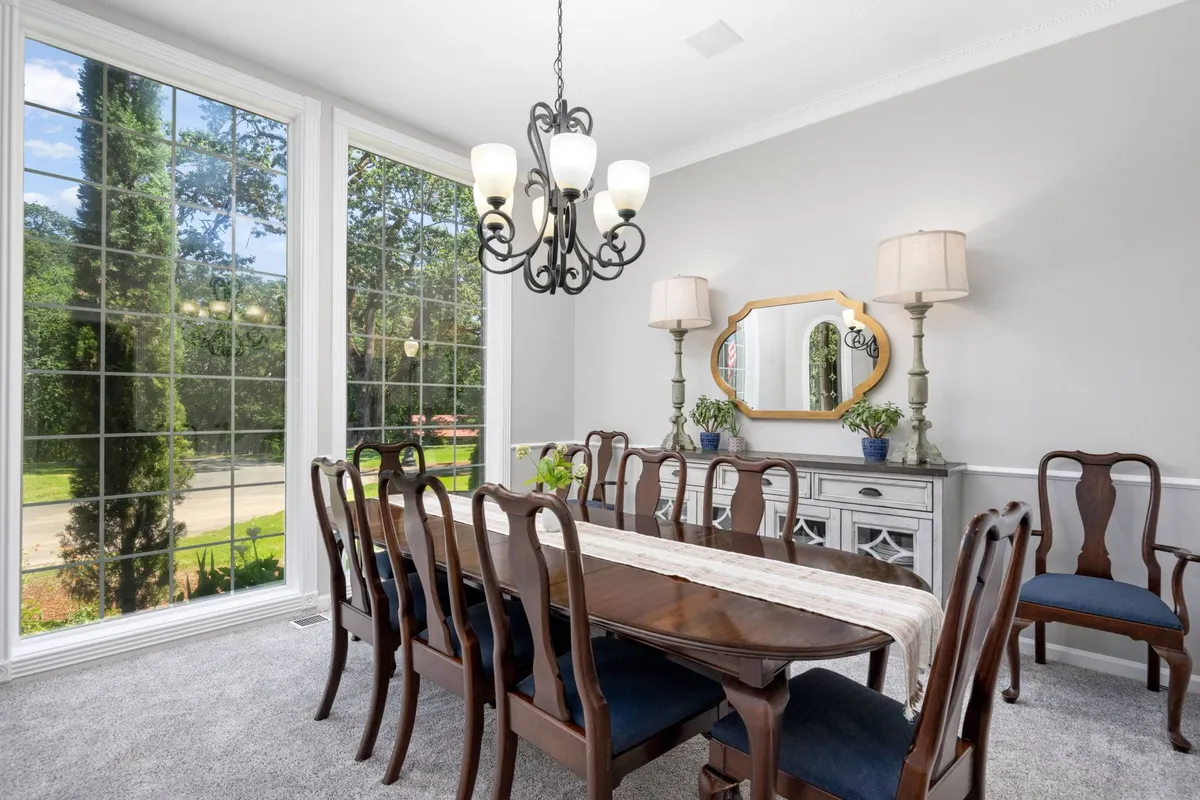 An elegant formal dining area with a grand table, chandelier, large windows offering garden views, sideboard, and decorative mirror. The space is designed for both intimate family dinners and celebratory gatherings.