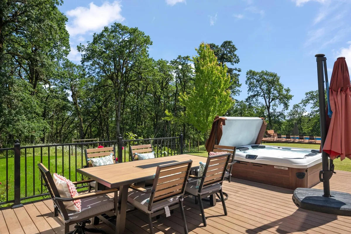 A serene outdoor dining table scene surrounded by lush trees, an inviting hot tub, and an umbrella, capturing an intimate area for relaxation and nature immersion. The environment is tranquil and features a blend of natural and landscaped elements.