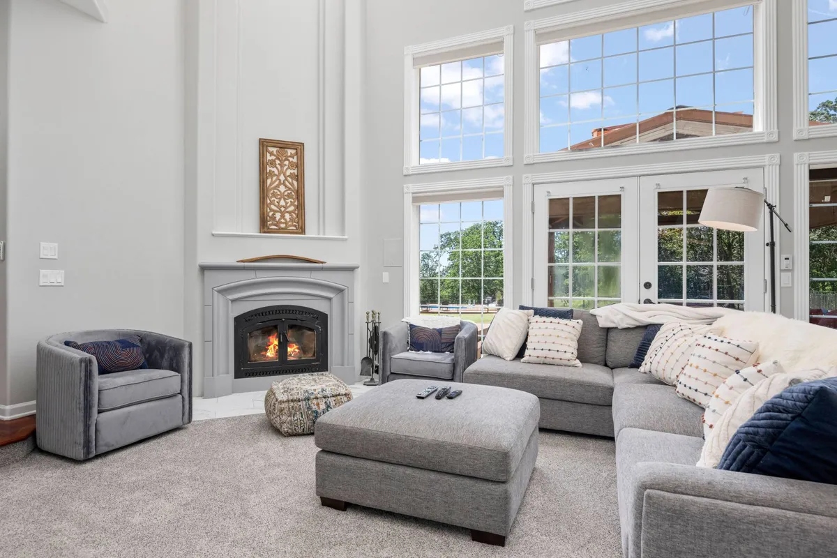 A cozy living area featuring a fireplace, comfortable seating, and expansive windows that overlook lush gardens and greenery. The large windows create a strong visual connection to the outdoor environment, alluding to the estate's natural surroundings and inviting atmosphere.