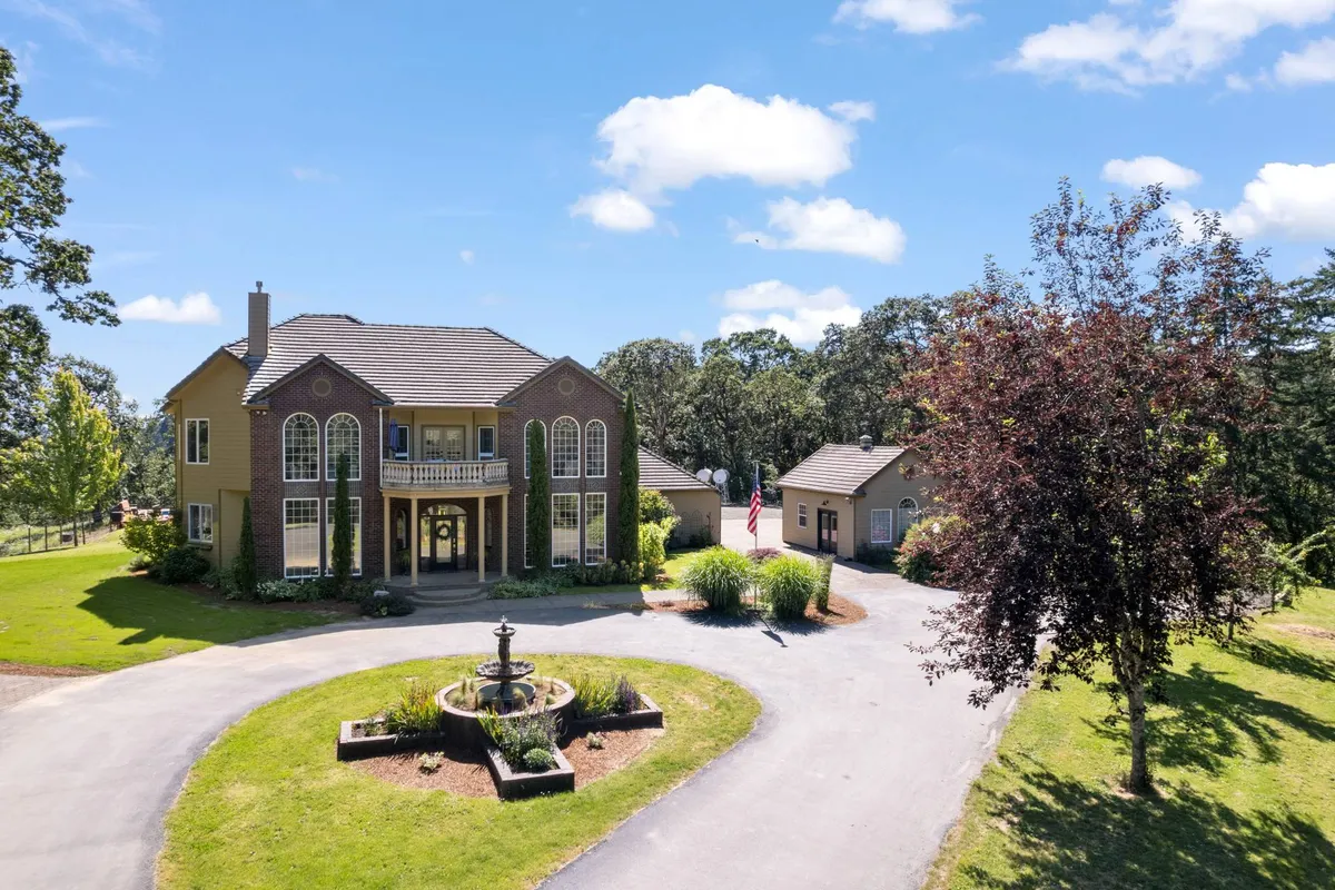 The image showcases the grand entrance of Oak Rise, featuring stately architecture framed by a lush canopy of Savannah oaks. A tranquil fountain is set amidst manicured landscaping and vibrant greenery, inviting guests into a serene and secluded oasis. The expansive front yard hints at ample space for outdoor gatherings and peaceful mornings alike, all surrounded by the natural beauty of the Pacific Northwest.